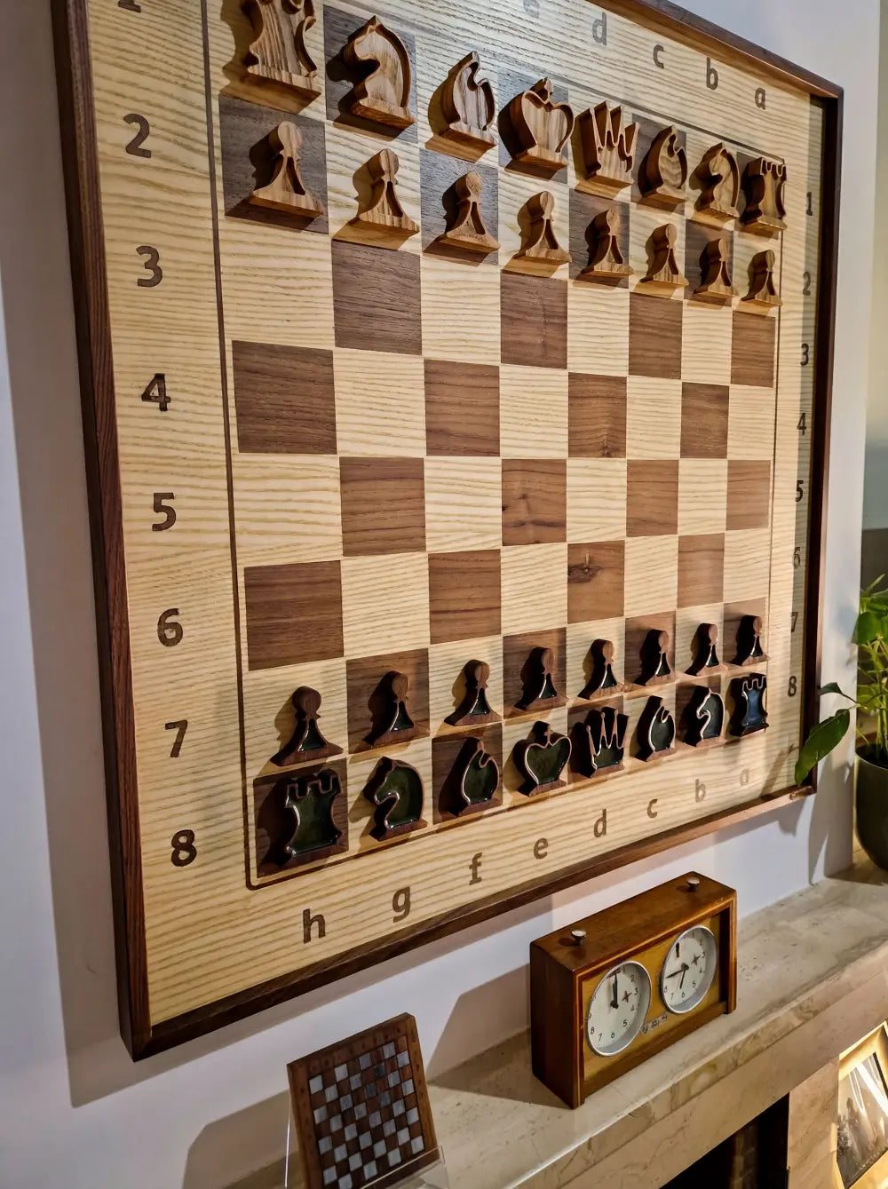 A large, vertical wooden chessboard hanging as wall art above a fireplace mantel, with a vintage wooden chess clock placed below it. Unique wall decor by ChessboArt.